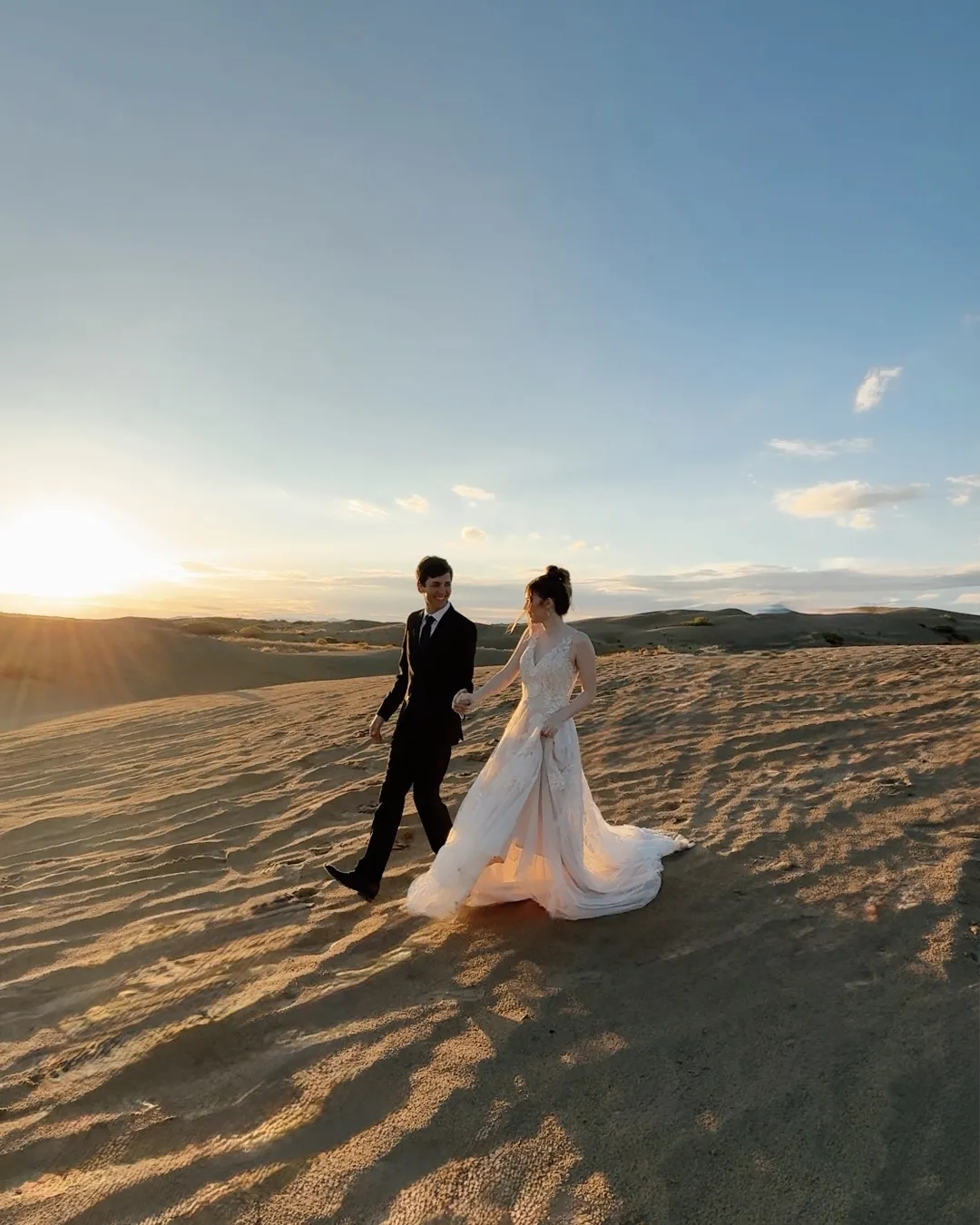 Bride and groom walking hand in hand across sand dunes at sunset.